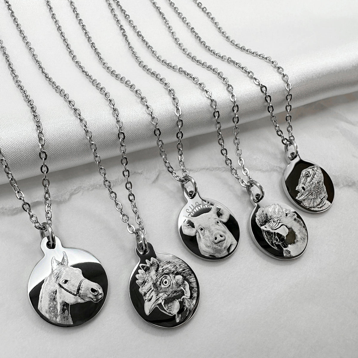 Five silver personalized pet portrait pendant necklaces with round engraved charms are arranged on a white satin fabric background. The charms depict different animals: horse, chicken, pig, parrot, and lizard or iguana.