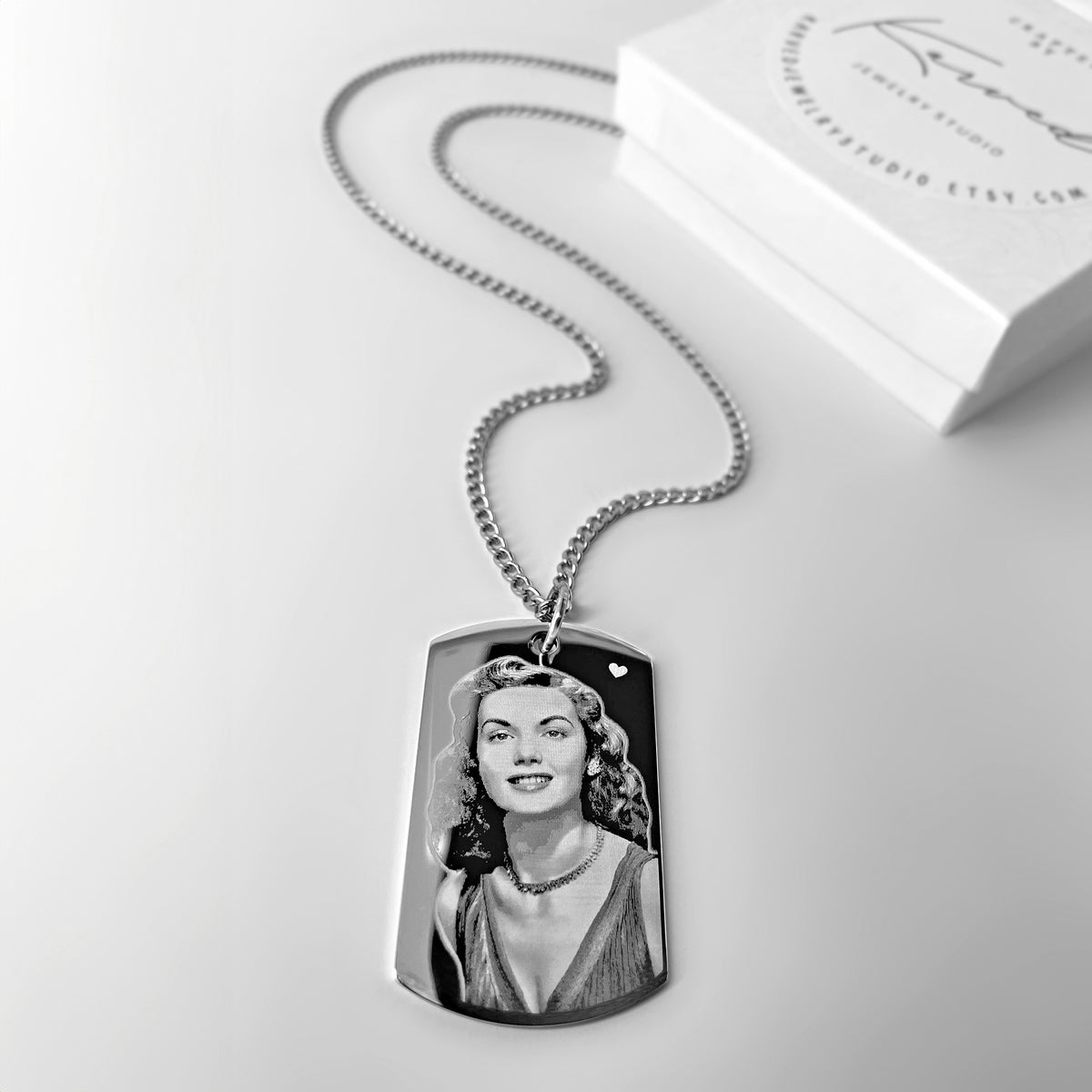 A silver custom dog tag necklace with an engraved personalized picture portrait pendant, displayed on a light gray surface with a branded jewelry box in the background.