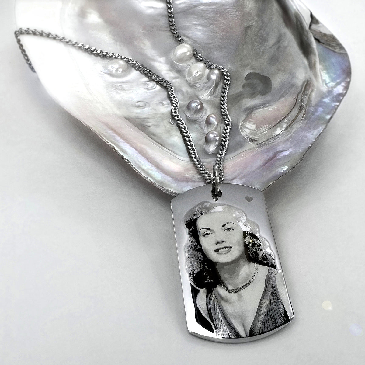 A silver pendant dog tag necklace displayed on a large iridescent shell. The pendant is a polished stainless steel dog tag photo portrait engraving of a woman, suspended from a chain with small pearl-like beads.
