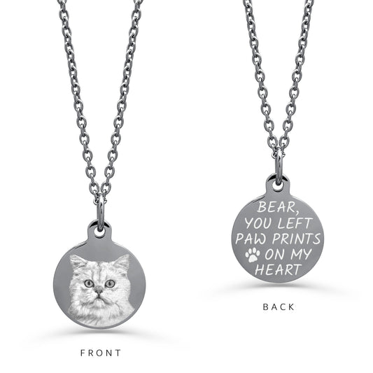 A silver round pendant necklace shown from both front and back on a white background. The front side features an engraved personalized portrait of a fluffy cat, and the back side is engraved with the memorial design 'YOU LEFT PAW PRINTS ON MY HEART' with a paw print.