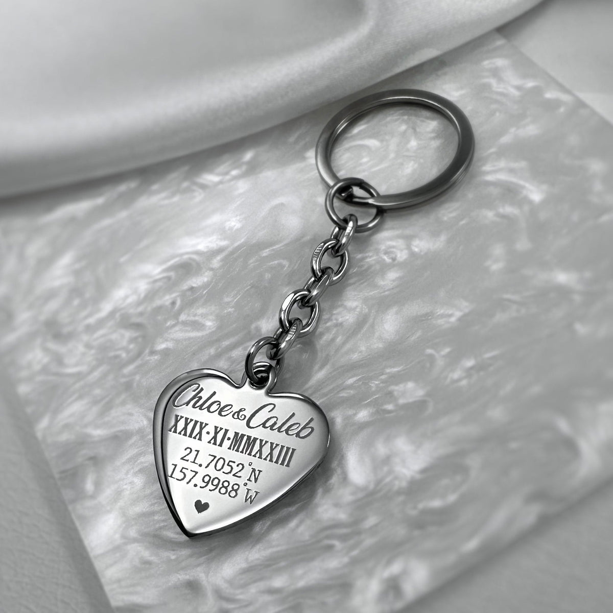 A silver heart-shaped keychain engraved with custom names, geographical coordinates, and personalized Roman numerals date displayed on a clean pearlescent background.