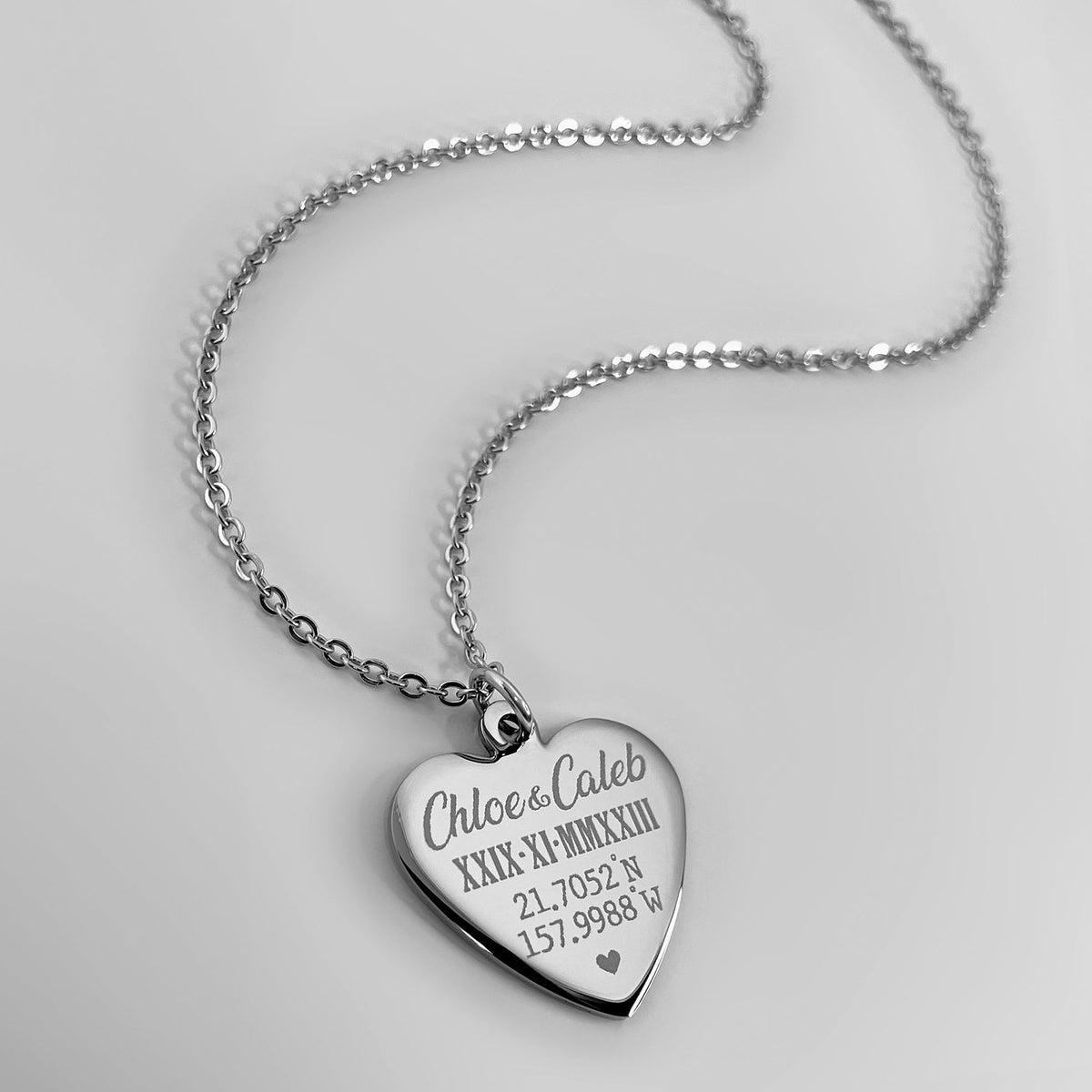 A silver heart-shaped pendant necklace engraved with custom names, geographical coordinates, and personalized Roman numerals date displayed on a clean light gray background.