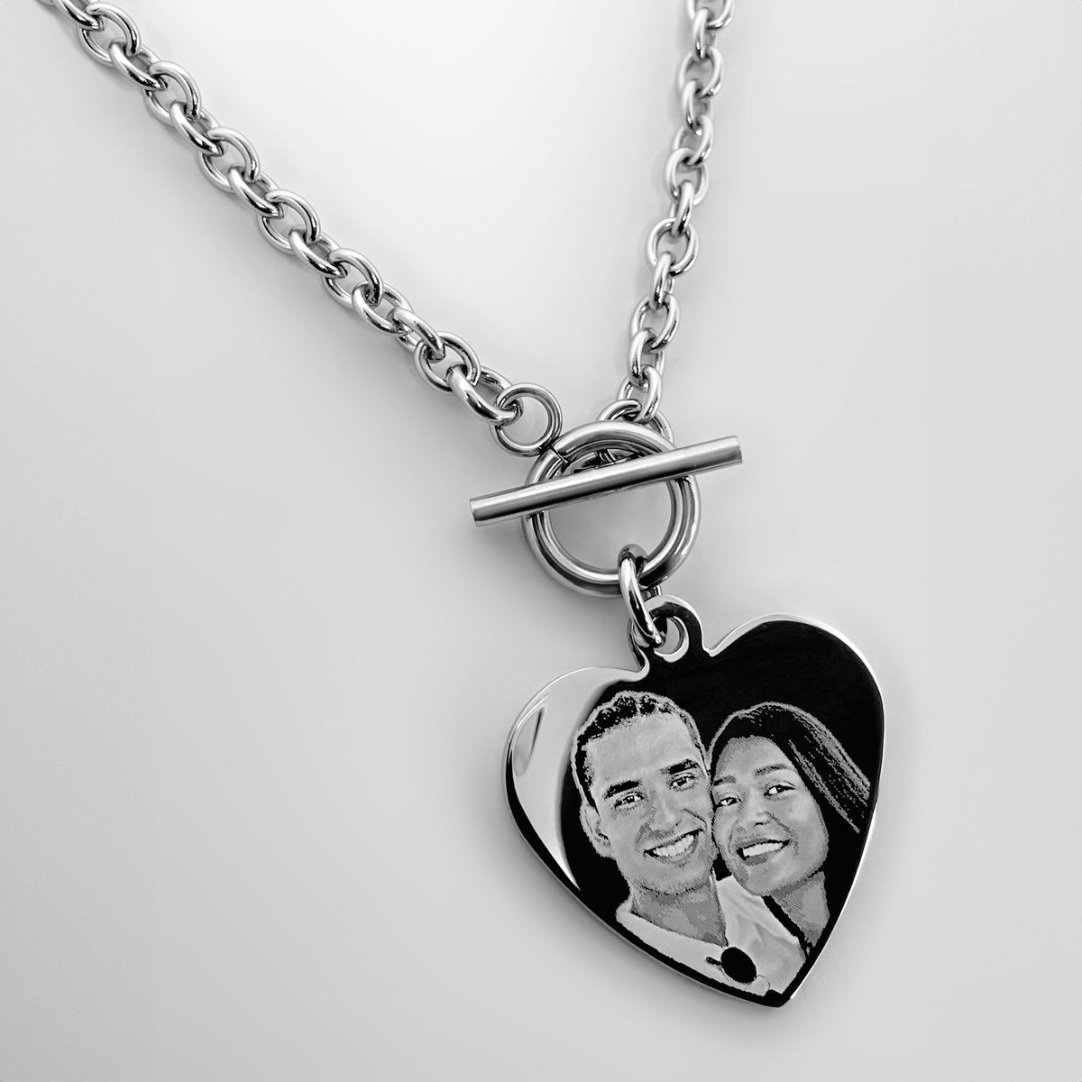 A silver chain necklace with a toggle clasp and a heart-shaped pendant. The heart pendant is engraved with a personalized photo of a couple.