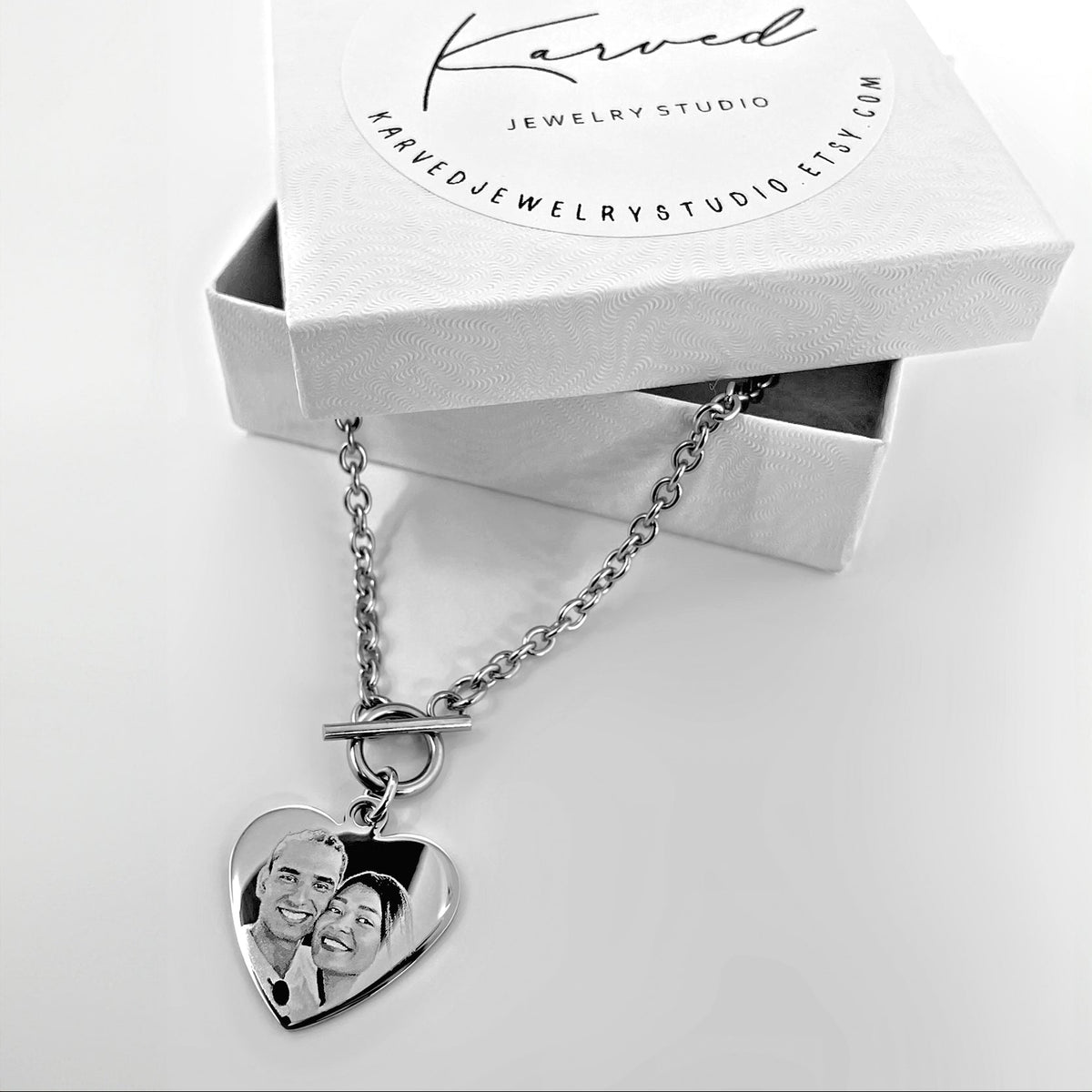 A silver heart pendant necklace with a custom photo engraving, displayed in front of its white Karved Jewelry Studio gift box.
