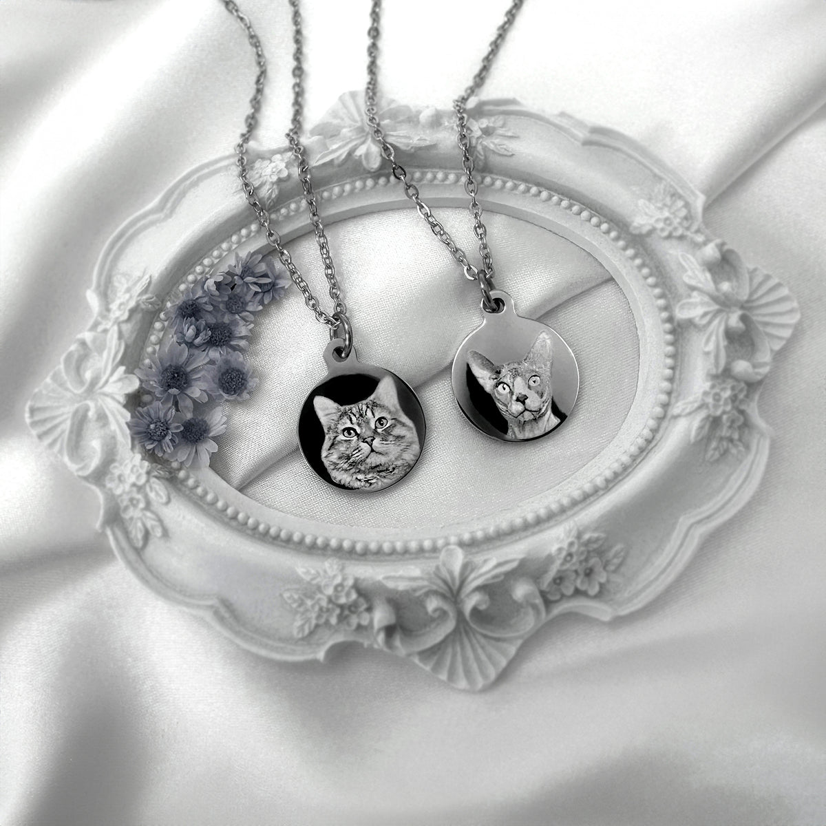 Two silver personalized cat portrait necklaces with round pendants are displayed on a white ornate oval tray with white satin fabric underneath.