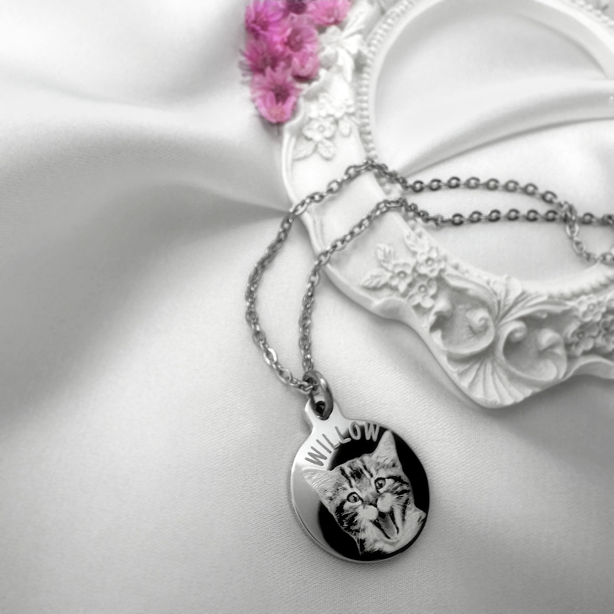 A silver necklace with a round engraved pendant featuring a personalized cat portrait and a custom name, styled on white satin fabric next to a white ornate dish and pink flowers.