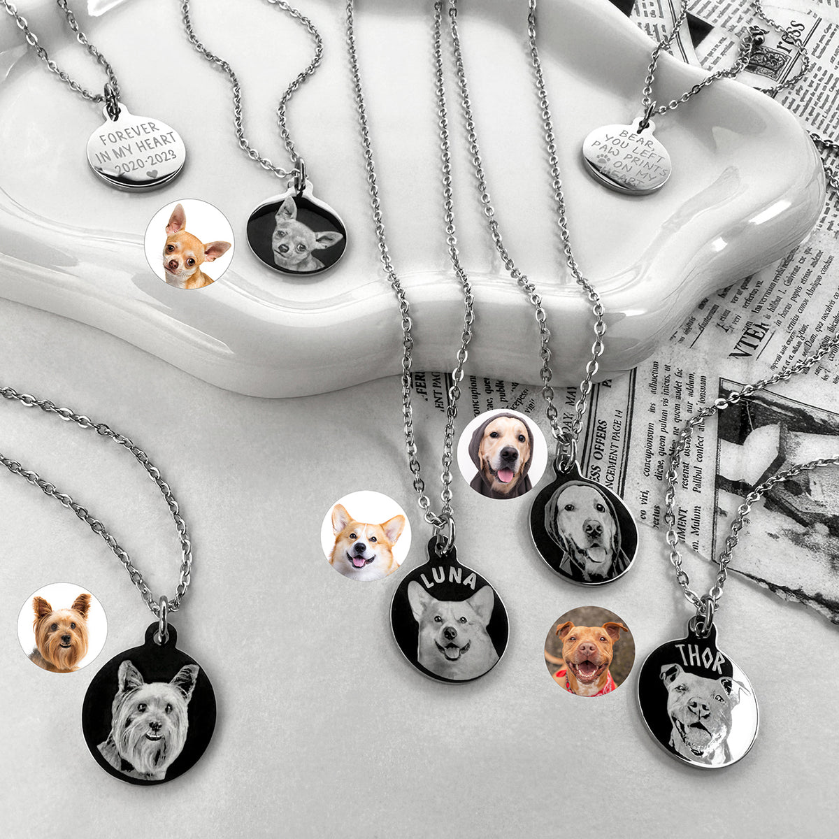 A styled flat-lay of multiple silver pet memorial necklaces with round pendants showing different dog portraits, displayed on a white ceramic dish and newspaper background.