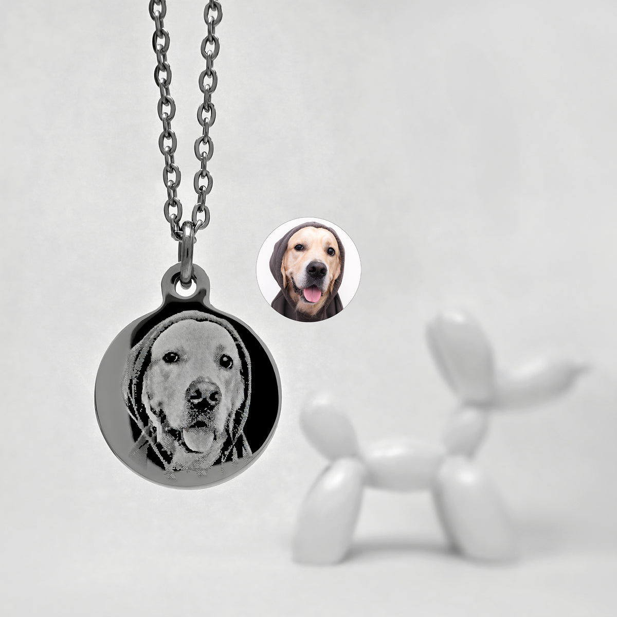 A round pendant necklace on a chain, featuring a personalized engraved dog portrait. A small circular inset shows the original dog photo used for the engraving, and a blurred white balloon-dog sculpture appears in the background.