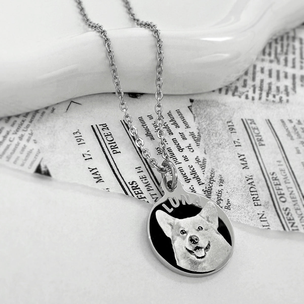 A silver round pendant necklace featuring a custom engraved portrait of a dog, displayed on a white ceramic dish over vintage newspaper pages.