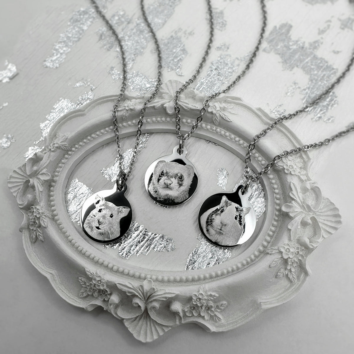 Three silver round pendant necklaces with custom pet portrait such as guinea pig, hamster and ferret charms resting on an ornate white decorative tray.