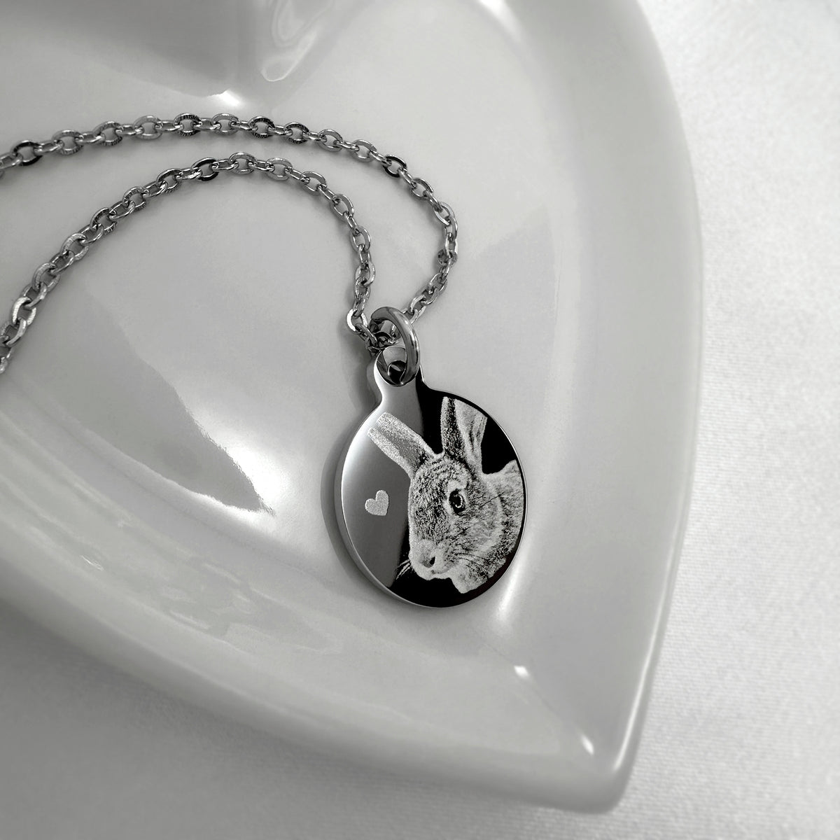 A silver necklace with a custom engraved round pendant featuring a personalized bunny rabbit portrait and a small white heart, displayed on a glossy white ceramic dish.
