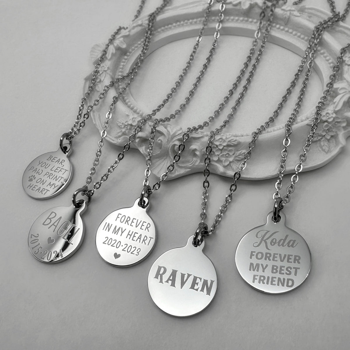 Four silver personalized round pendant necklaces with chains, displayed on a white decorative tray. The pendants are engraved with different pet designs and personalized memorial messages.