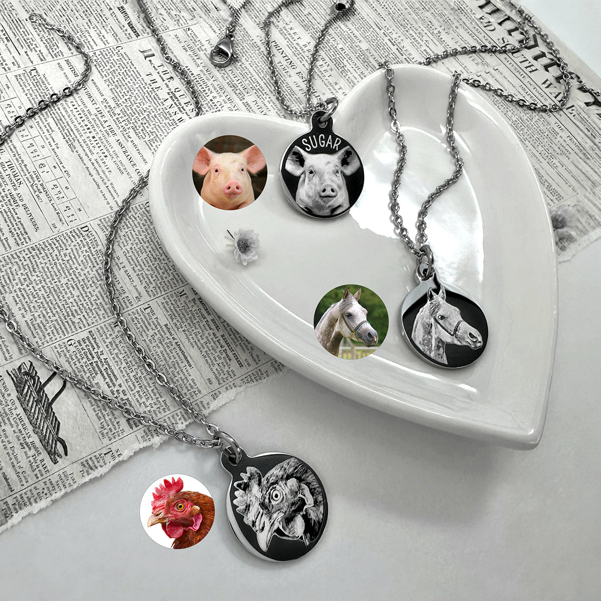 Three silver chain necklaces with round custom pet portrait pendants are displayed on a white heart-shaped dish and a newspaper background. The pendants feature custom photos of a pig, a horse, and a chicken, with one pendant showing a personalized horse portrait with a custom name.