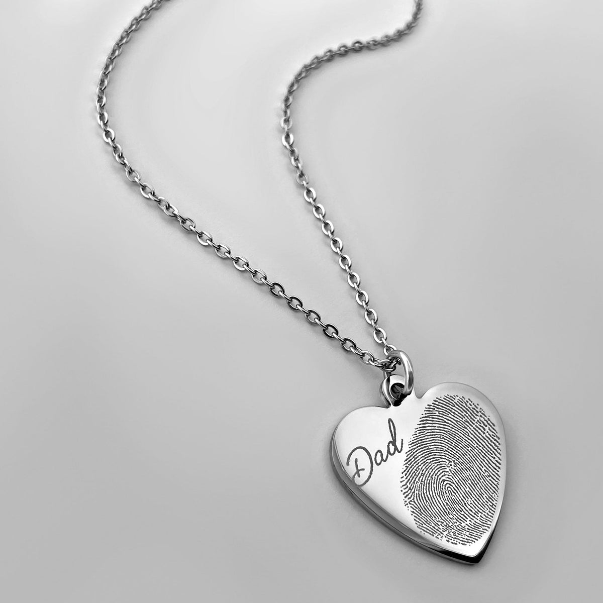 A silver heart-shaped keepsake pendant necklace engraved with the handwritten word 'Dad' and a custom fingerprint, shown on a matching silver chain against a light gray background.