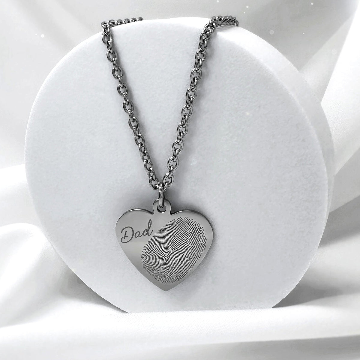 A silver heart pendant necklace with a custom engraved fingerprint and handwritten word 'Dad' engraved on the front, displayed on a white circular stand.