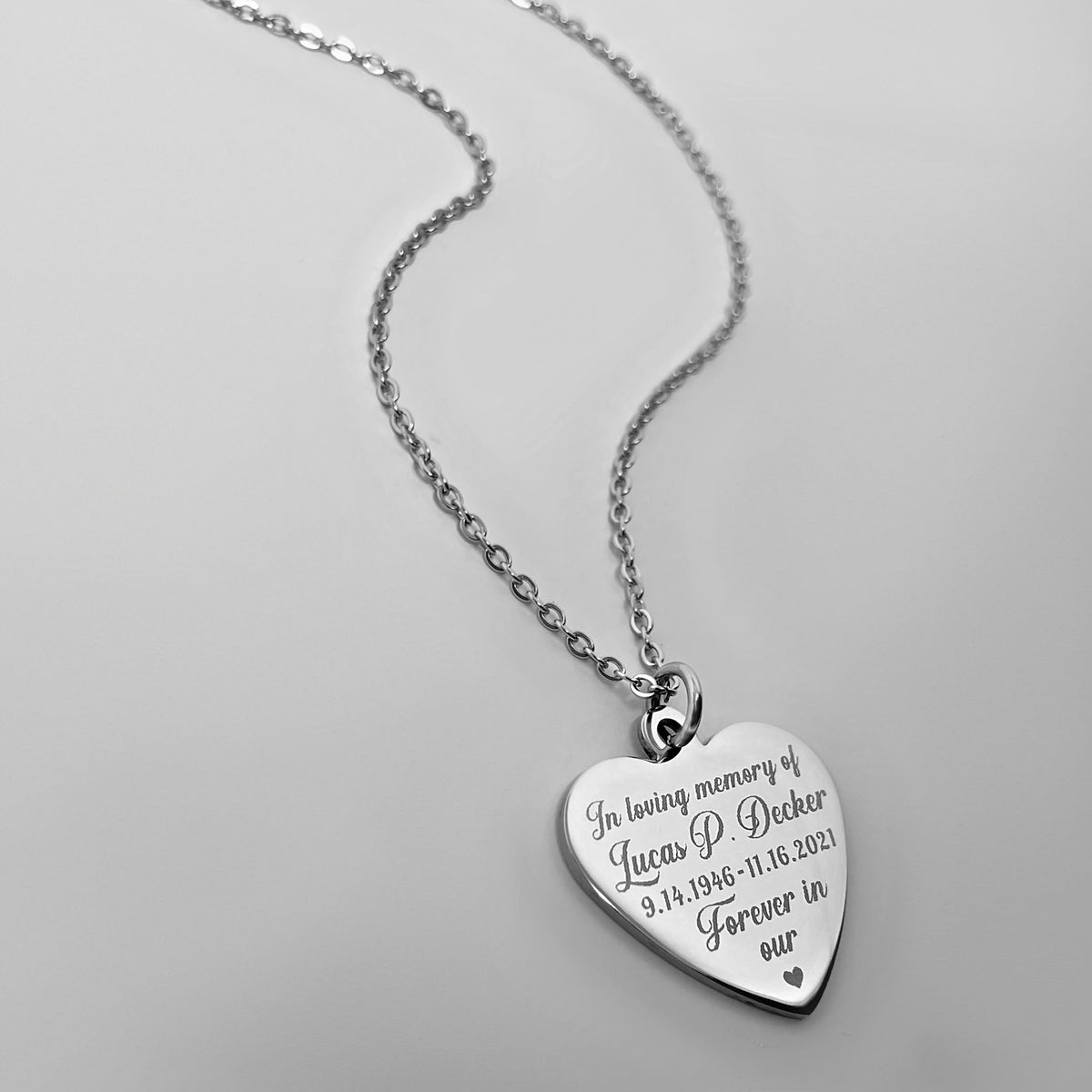 A silver heart-shaped pendant necklace engraved with personalized name, DOB-DOD dates and a custom memorial saying.