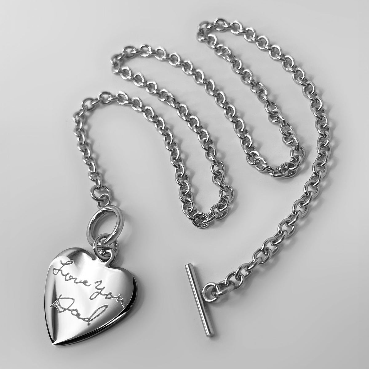 A silver heart-shaped pendant necklace engraved with person's actual handwriting, displayed with a matching chain with a toggle clasp against a light gray background.