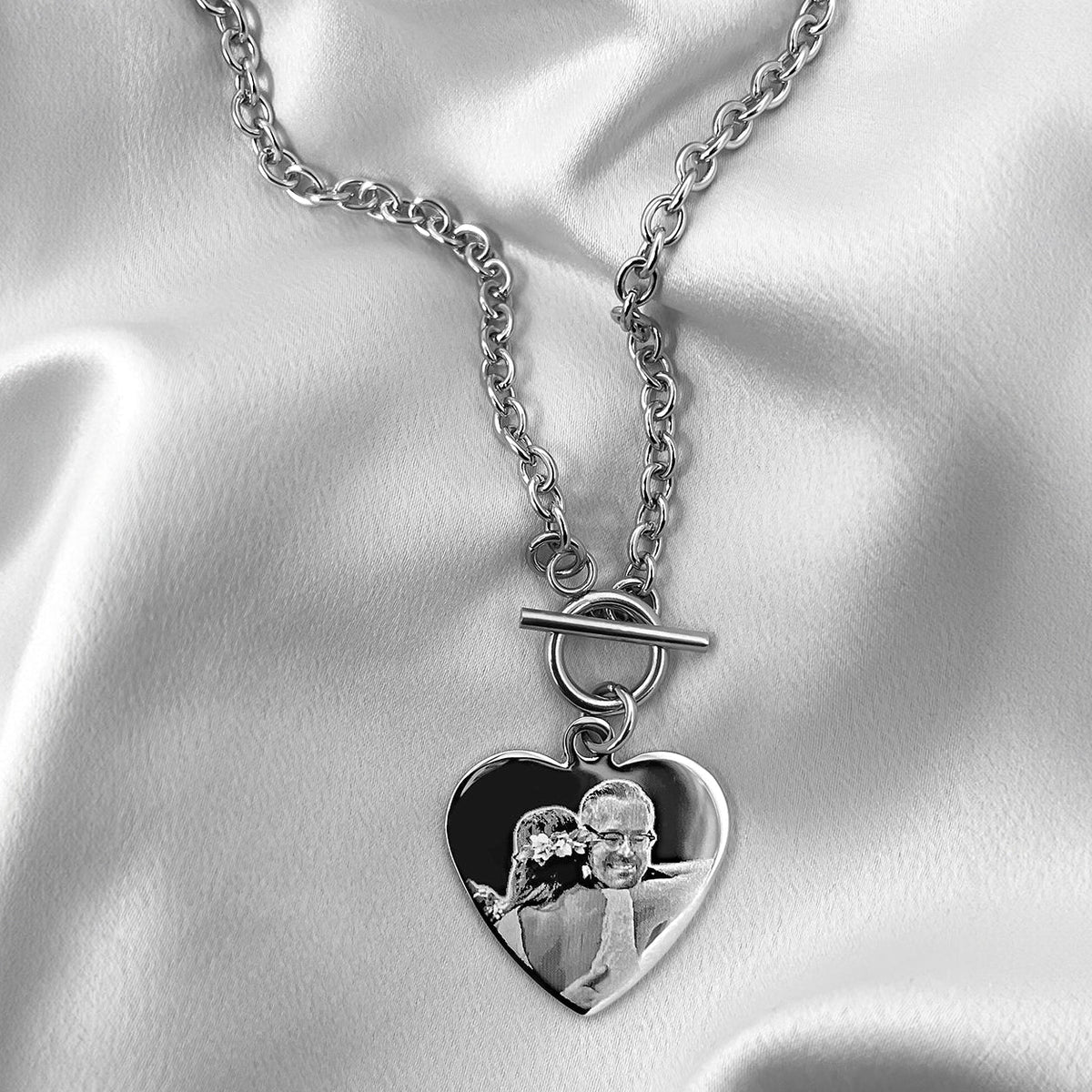 A silver heart pendant choker with a toggle clasp chain with a personalized photo charm, displayed on a draped white satin fabric background.