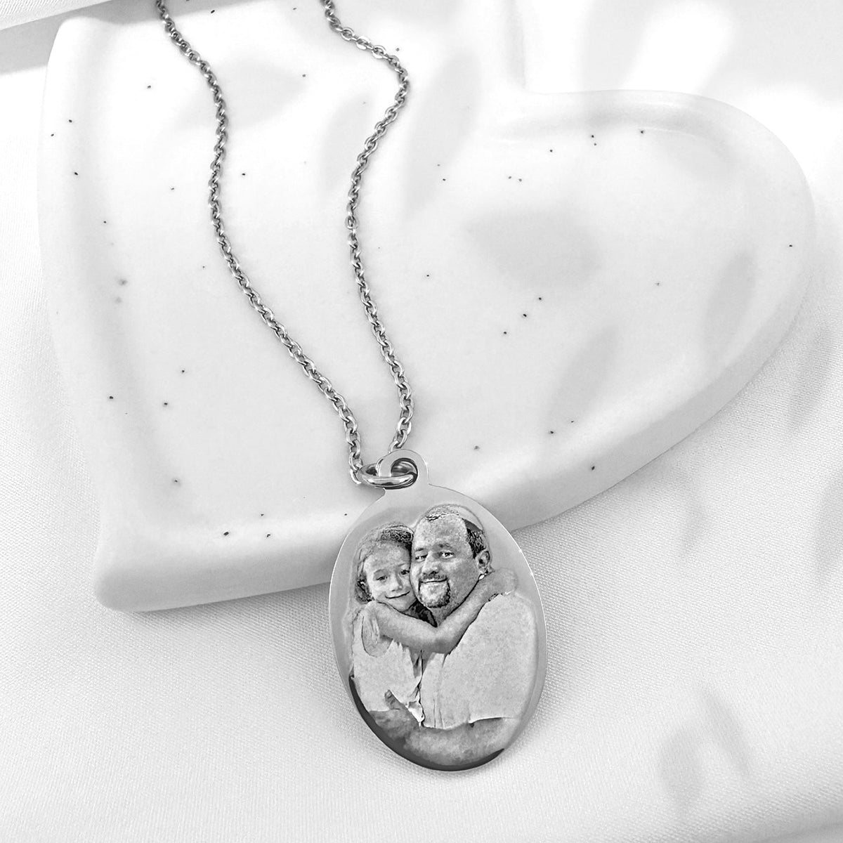 A silver oval pendant necklace featuring a custom portrait photo of a father and daughter, displayed on a white speckled heart-shaped tray.