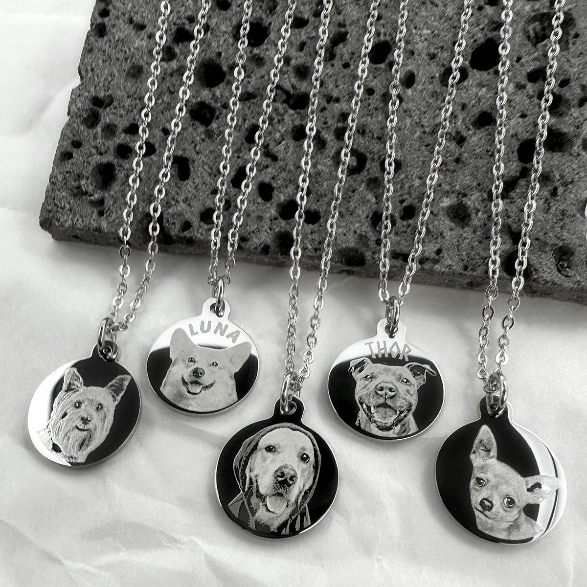 Five silver personalized pet portrait necklaces with round engraved pendants showing different dog faces, displayed on a white fabric background with a dark stone slab.