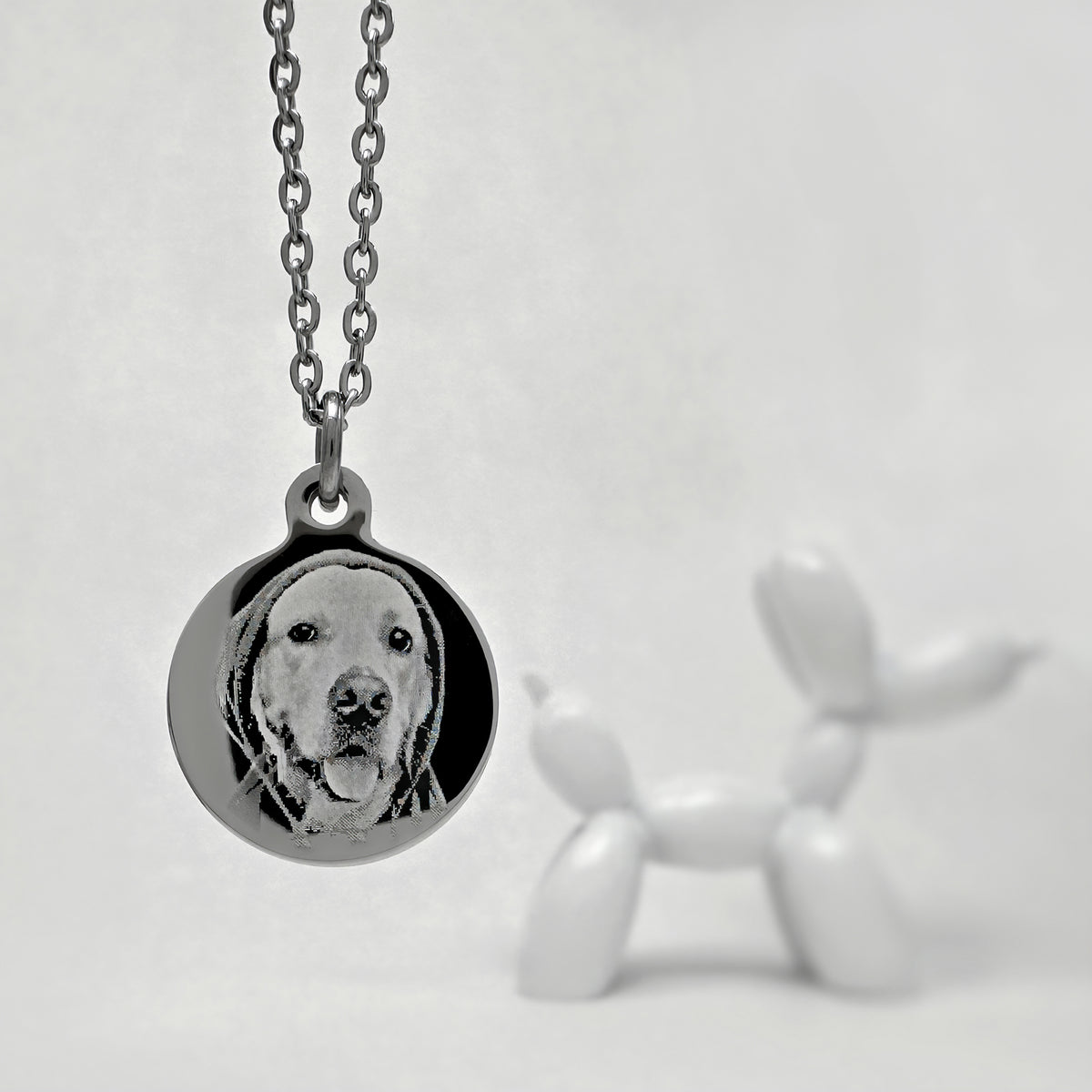 A silver round pendant necklace featuring a custom portrait of a dog, shown hanging from a silver chain against a light gray background with a blurred white balloon dog figurine in the foreground.