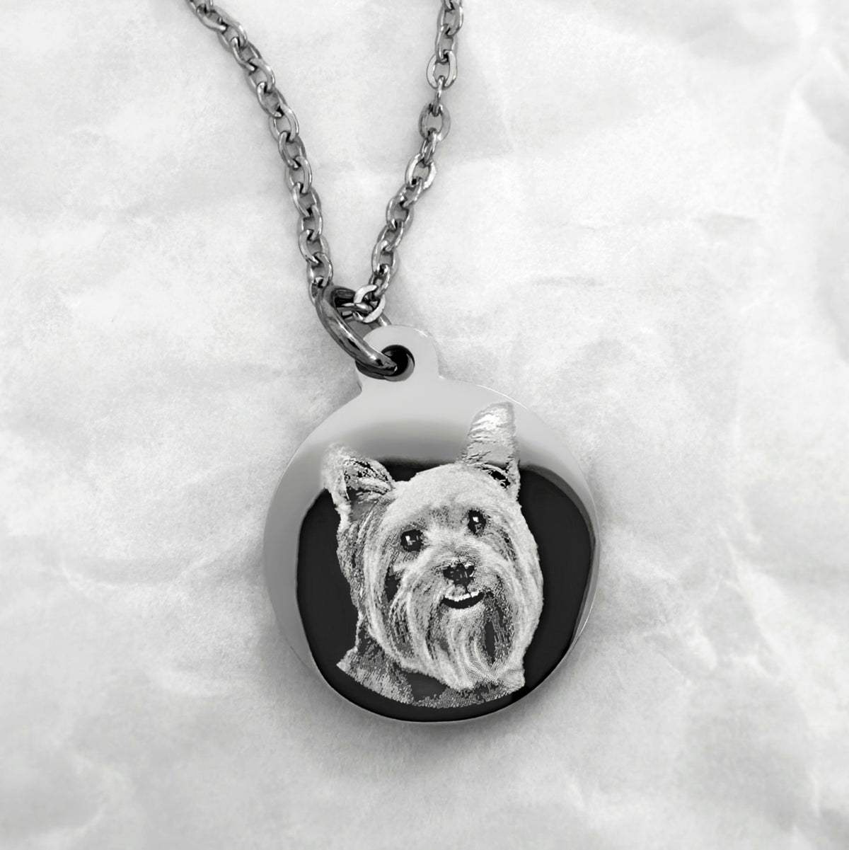 A silver round personalized pendant necklace featuring a custom portrait of a yorkie, displayed on a soft white textured fabric background.