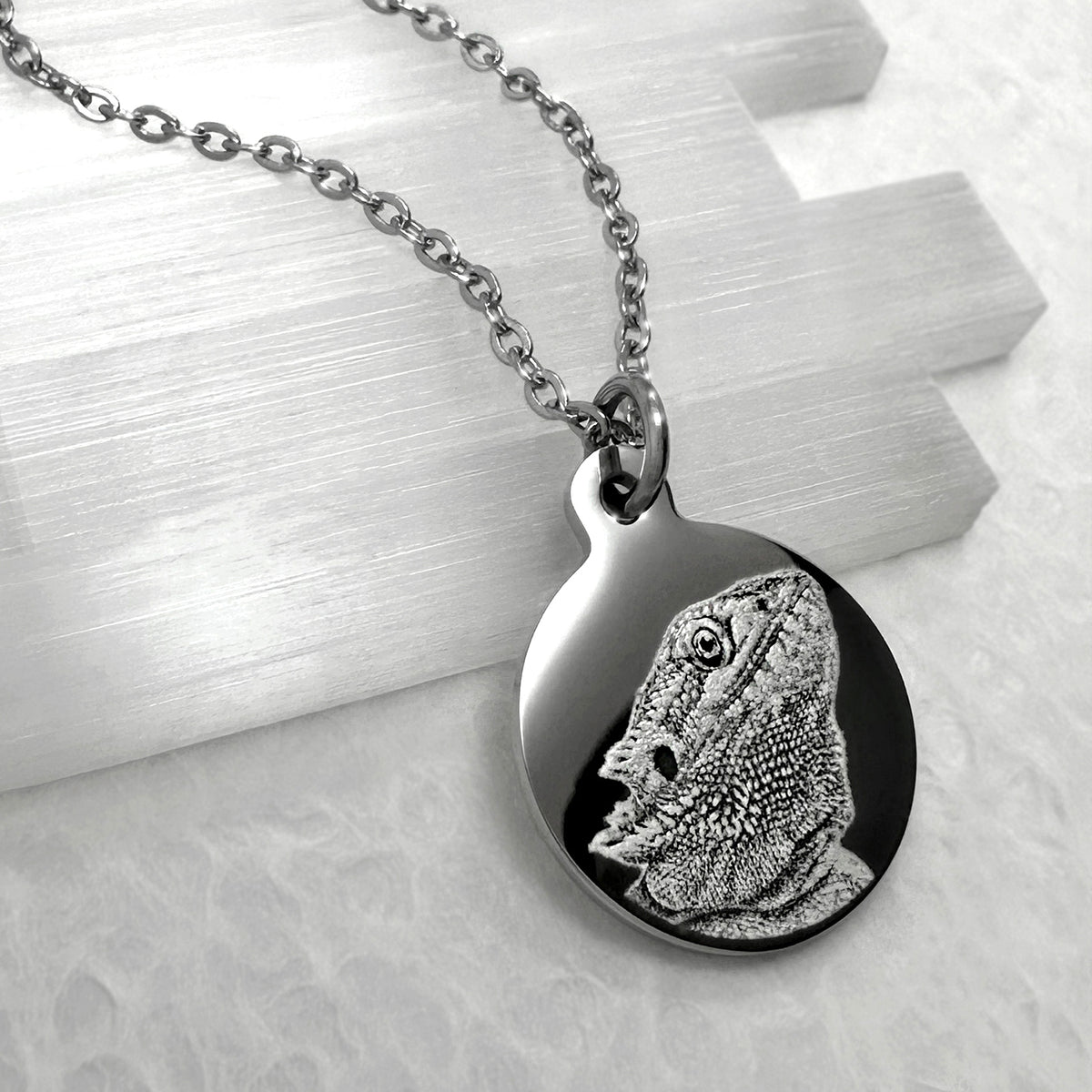 A silver round pendant necklace featuring an engraved personalized bearded dragon, displayed on a textured white surface with a brushed metal block. 