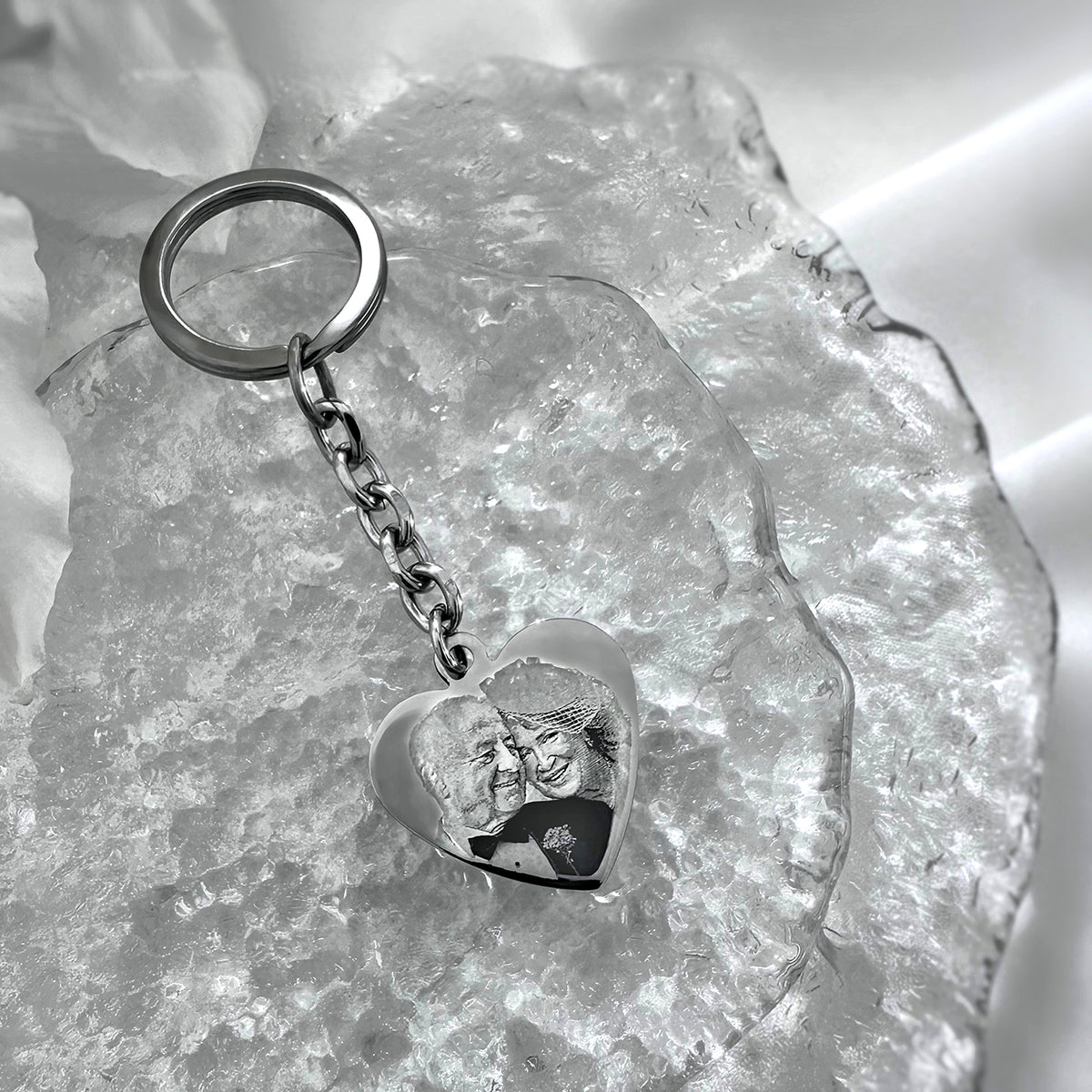 A silver heart-shaped keychain with a custom photo engraving, resting on a textured clear crystal slab.