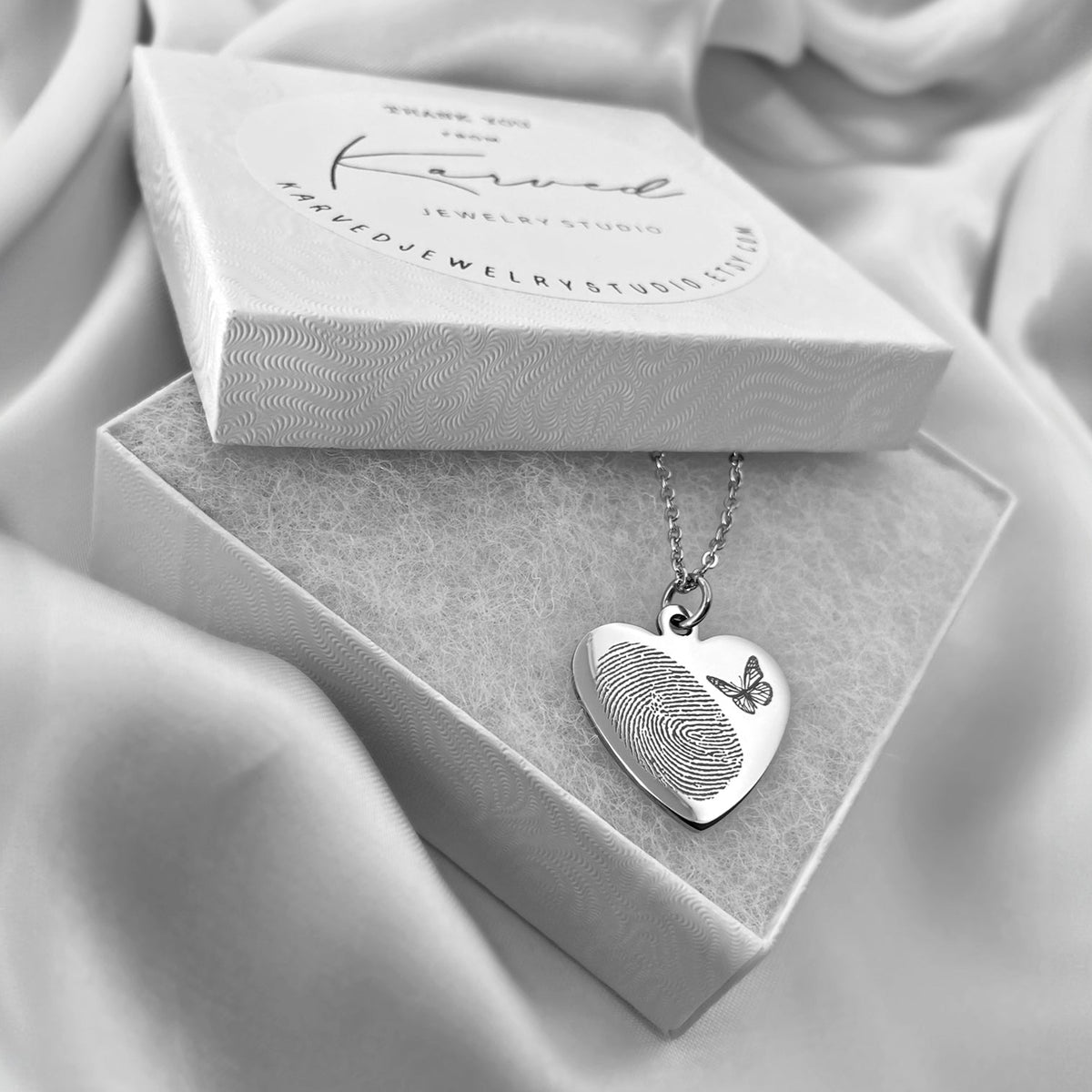 A silver heart-shaped pendant necklace with an actual engraved fingerprint and butterfly design, displayed in an open jewelry box on draped satin fabric.