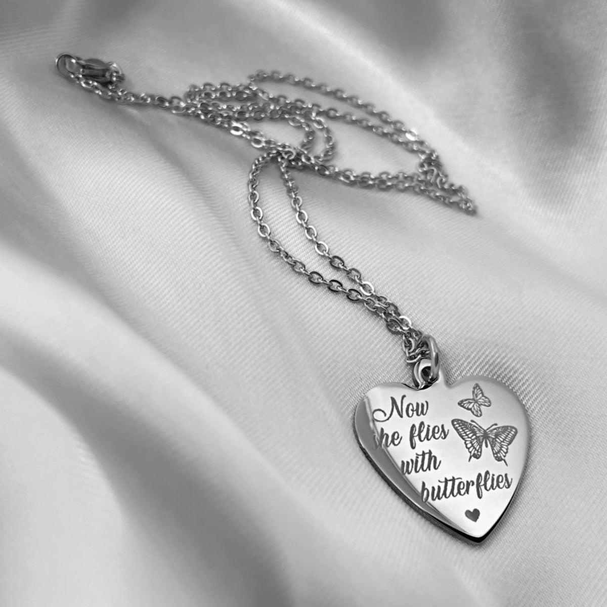A stainless-steel heart-shaped pendant necklace with the engraved custom memorial poem 'Now she flies with butterflies' and butterfly design, displayed on a draped white satin fabric background.