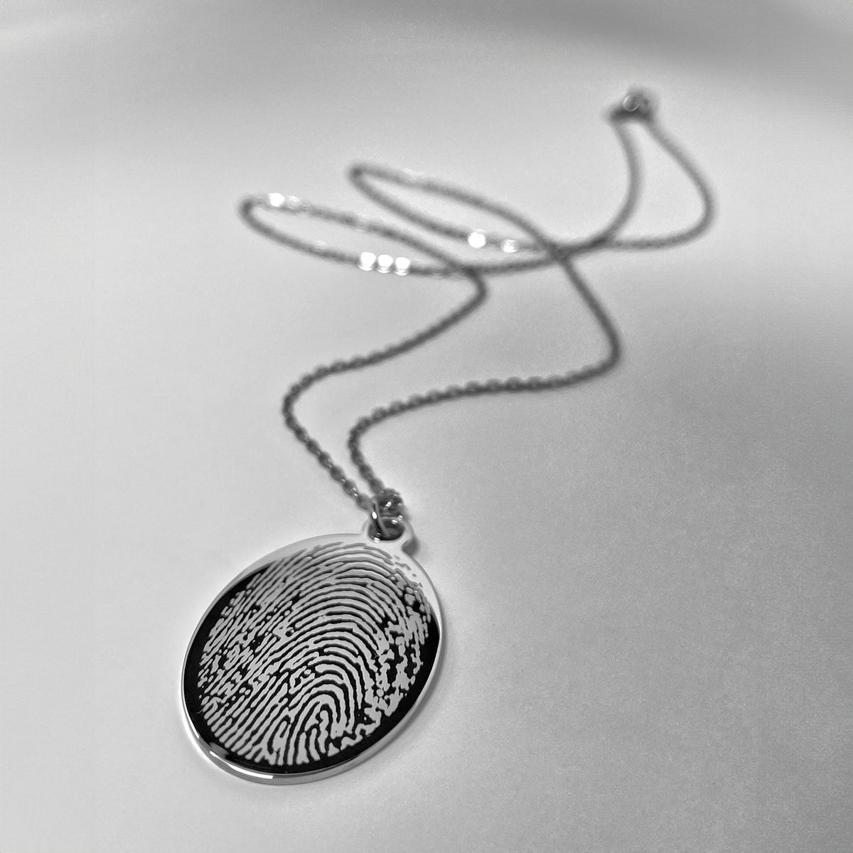 A stainless-steel oval pendant necklace engraved with a custom fingerprint design, resting on a light gray surface.