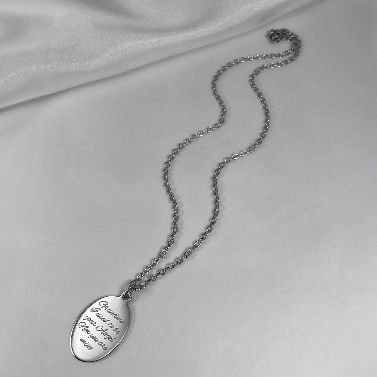 A stainless-steel oval pendant necklace with engraved personalized memorial saying displayed on a soft white satin fabric background.