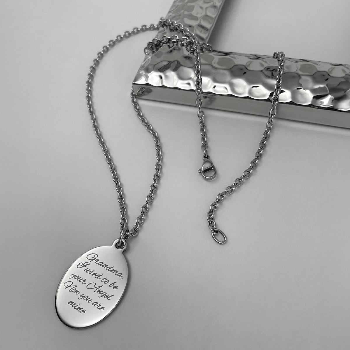 A silver oval pendant necklace engraved with the personalized memorial message 'Grandma, I just want to be your Angel, now you are mine', displayed on a textured silver stand against a light gray background.