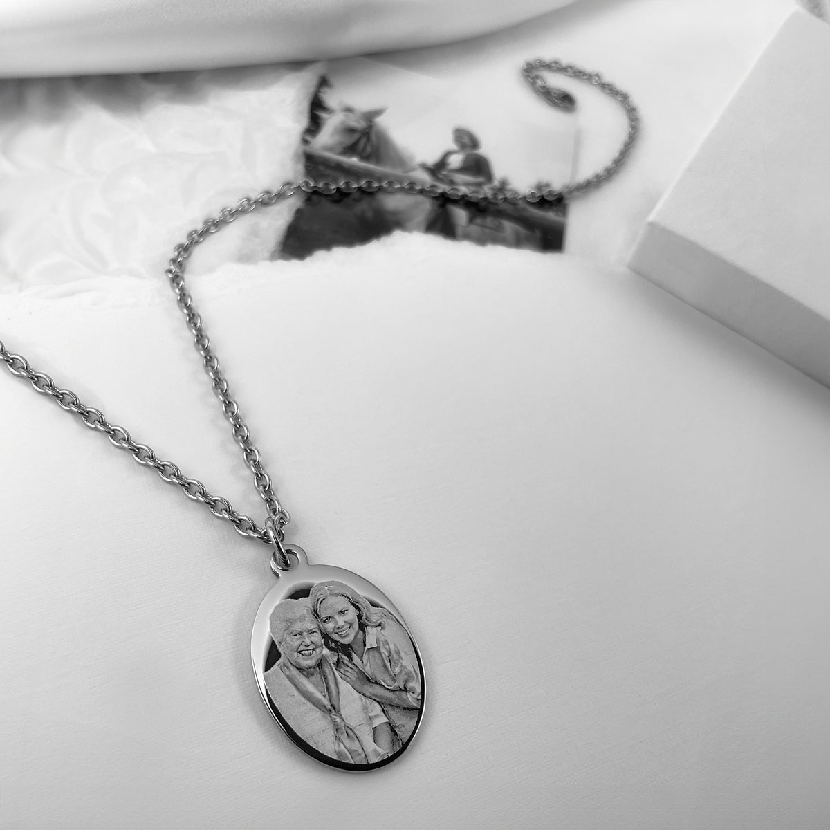 A silver oval pendant necklace engraved with a custom photo of a grandma and a granddaughter, displayed on a white textured fabric surface with a blurred photo card and gift box in the background.