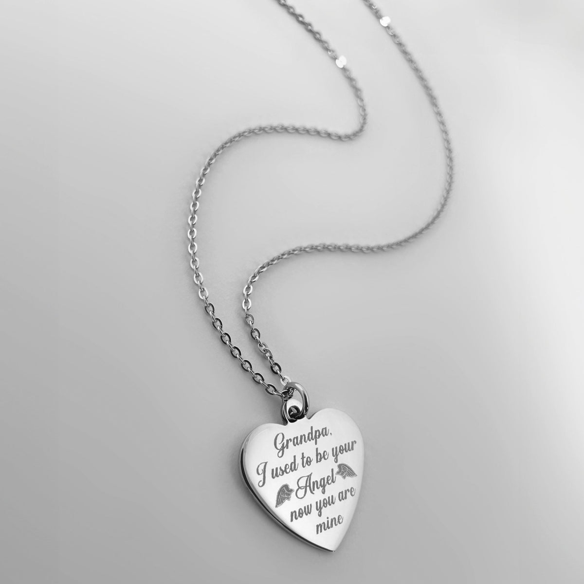 A silver heart-shaped pendant necklace engraved with the personalized message 'Grandpa, I used to be your Angel now you are mine' and small angel wings, shown on a light gray background.
