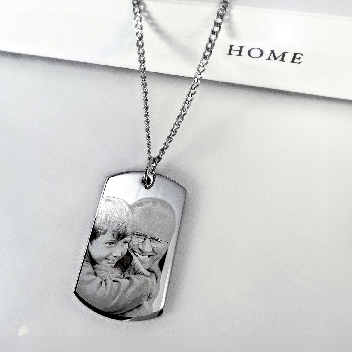 A silver dog tag necklace with a custom grandpa and grandson photo engraving, displayed on a white background with a book that reads 'HOME'.