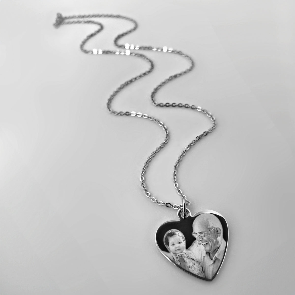 A silver heart-shaped pendant necklace featuring a custom photo of a grandpa and a grandchild, displayed on a light gray background.