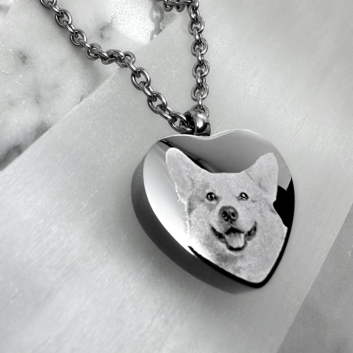 A heart-shaped urn pendant necklace with a polished metallic finish, featuring a custom engraved dog portrait. The necklace is displayed on a light marble surface.