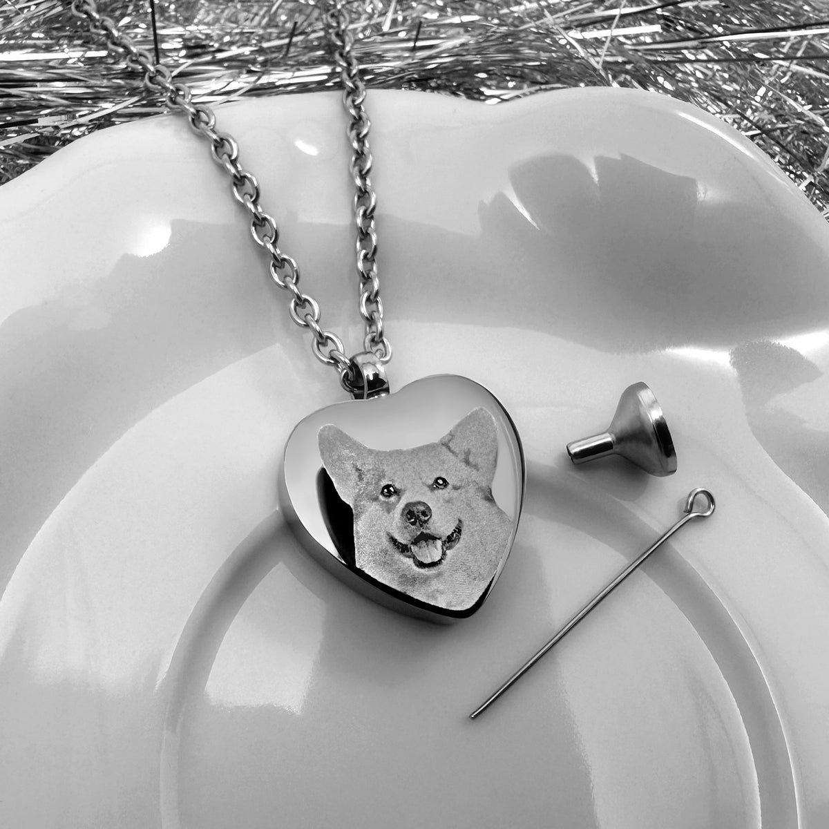 A silver heart-shaped pet urn necklace with a custom engraved dog portrait, displayed on a white ceramic dish alongside a fill kit, that consists of a funnel and a pin.