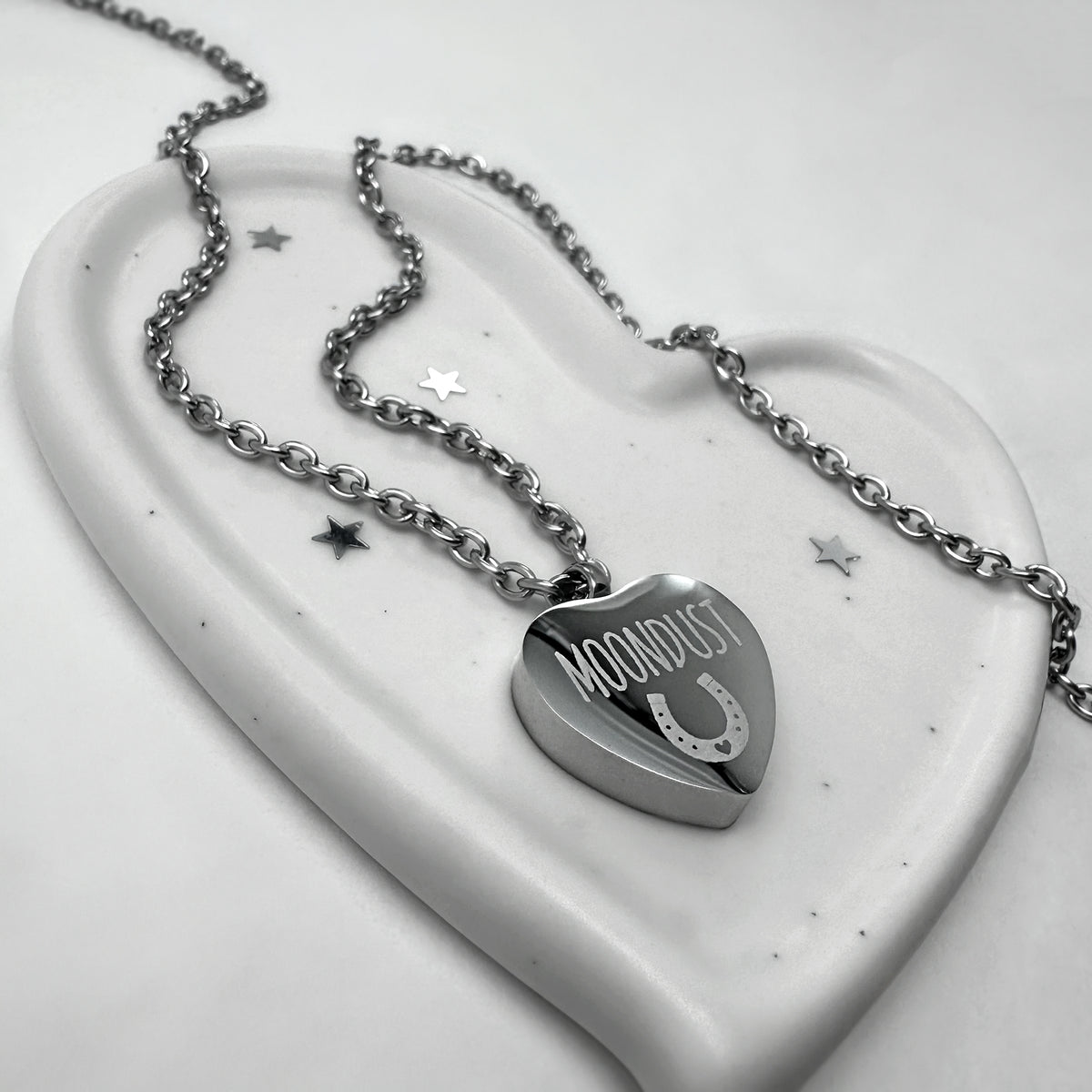 A stainless-steel heart-shaped urn pendant necklace engraved with the word a custom pet name and design, displayed on a white ceramic heart-shaped tray.