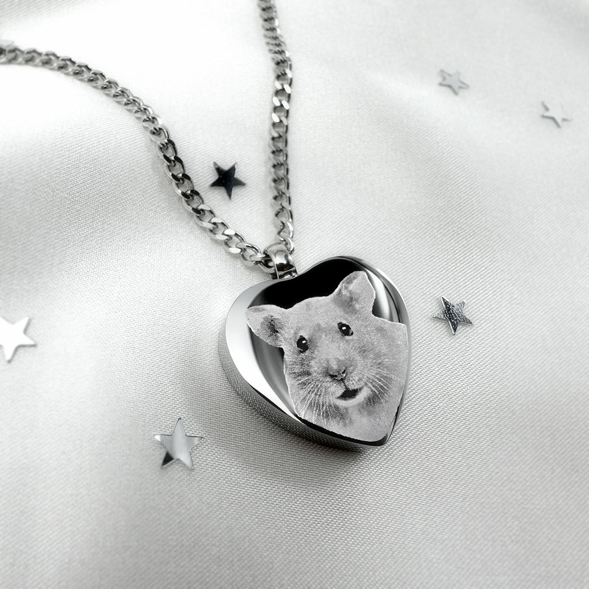A silver heart-shaped urn pendant necklace featuring a custom hamster portrait, displayed on a white satin fabric with scattered star-shaped confetti.