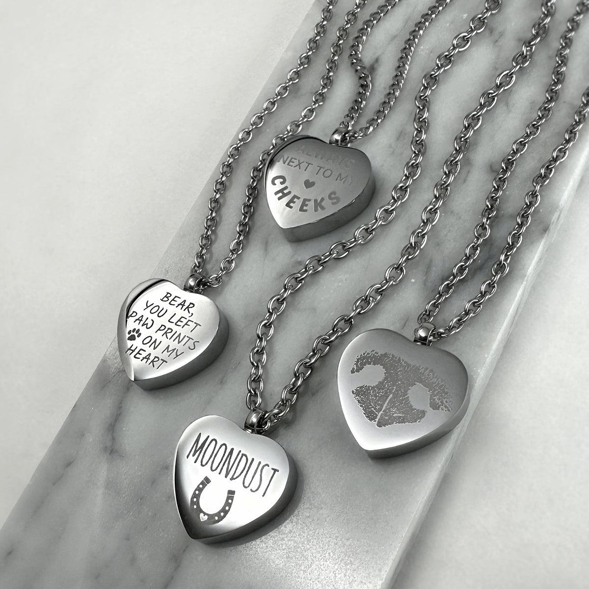 Four silver heart-shaped urn pendant necklaces arranged on a white marble tray. The pendants are engraved with memorial words and designs, including 'NEXT TO MY HEART', 'BEAR, YOU LEFT PAW PRINTS ON MY HEART', 'HORSE NAME', and actual nose print and paw print.