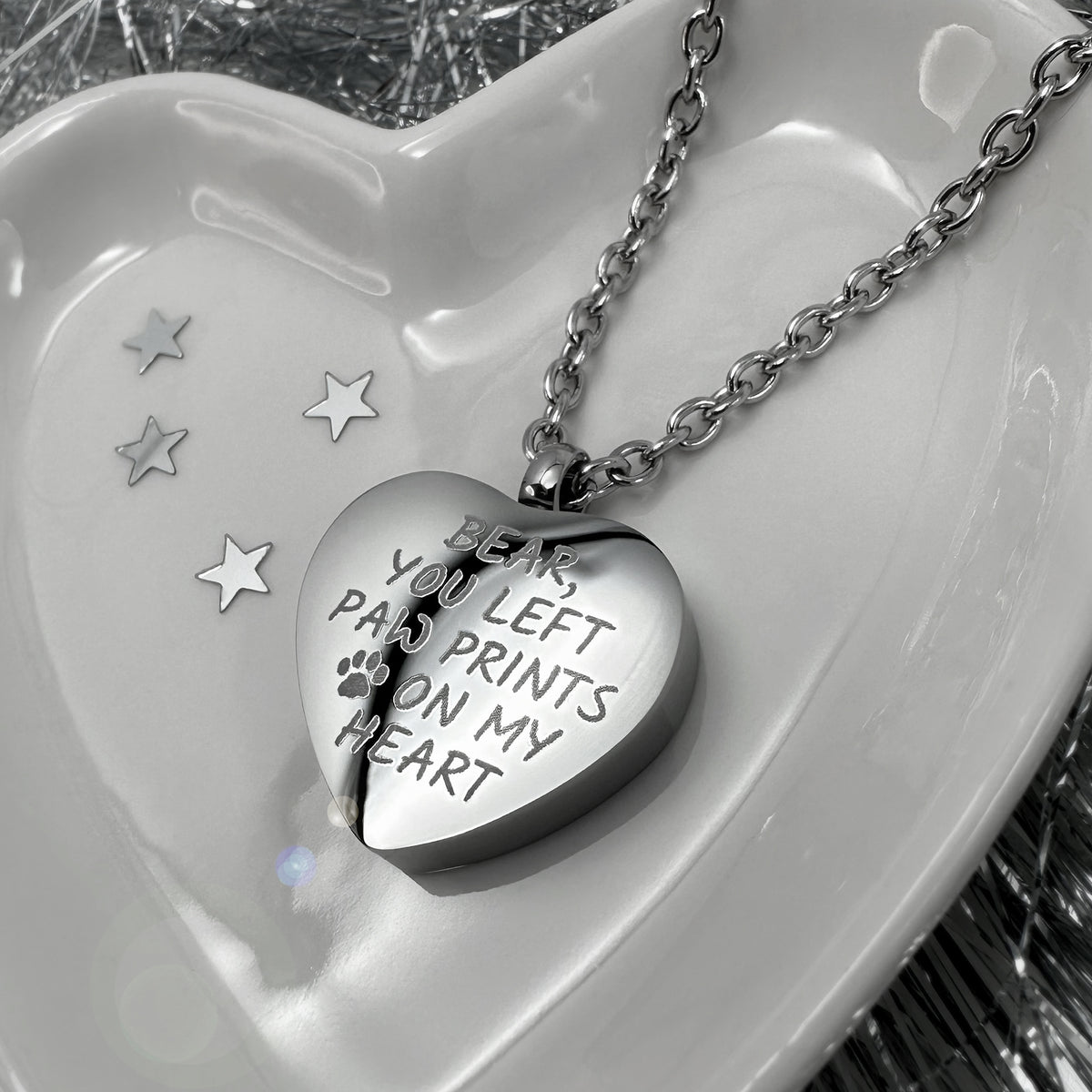 A stainless-steel heart-shaped urn pendant necklace engraved with 'BEAR, YOU LEFT PAW PRINTS ON MY HEART' and a paw print symbol, resting on a white ceramic heart-shaped dish.
