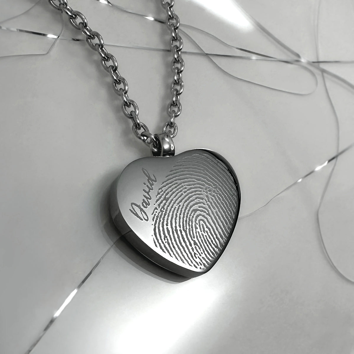 A silver heart-shaped urn pendant necklace engraved with a fingerprint pattern and a personalized name, resting on a glossy white surface.
