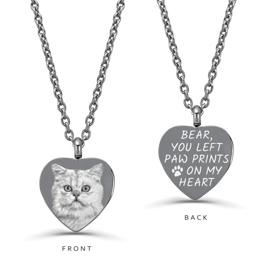 Heart-shaped urn pendant necklace on stainless steel chains, showcasing FRONT and BACK engraving. The left front pendant shows a grayscale photo of a cat, while the right back pendant is engraved with the text 'BEAR, YOU LEFT PAW PRINTS ON MY HEART' and a paw print.