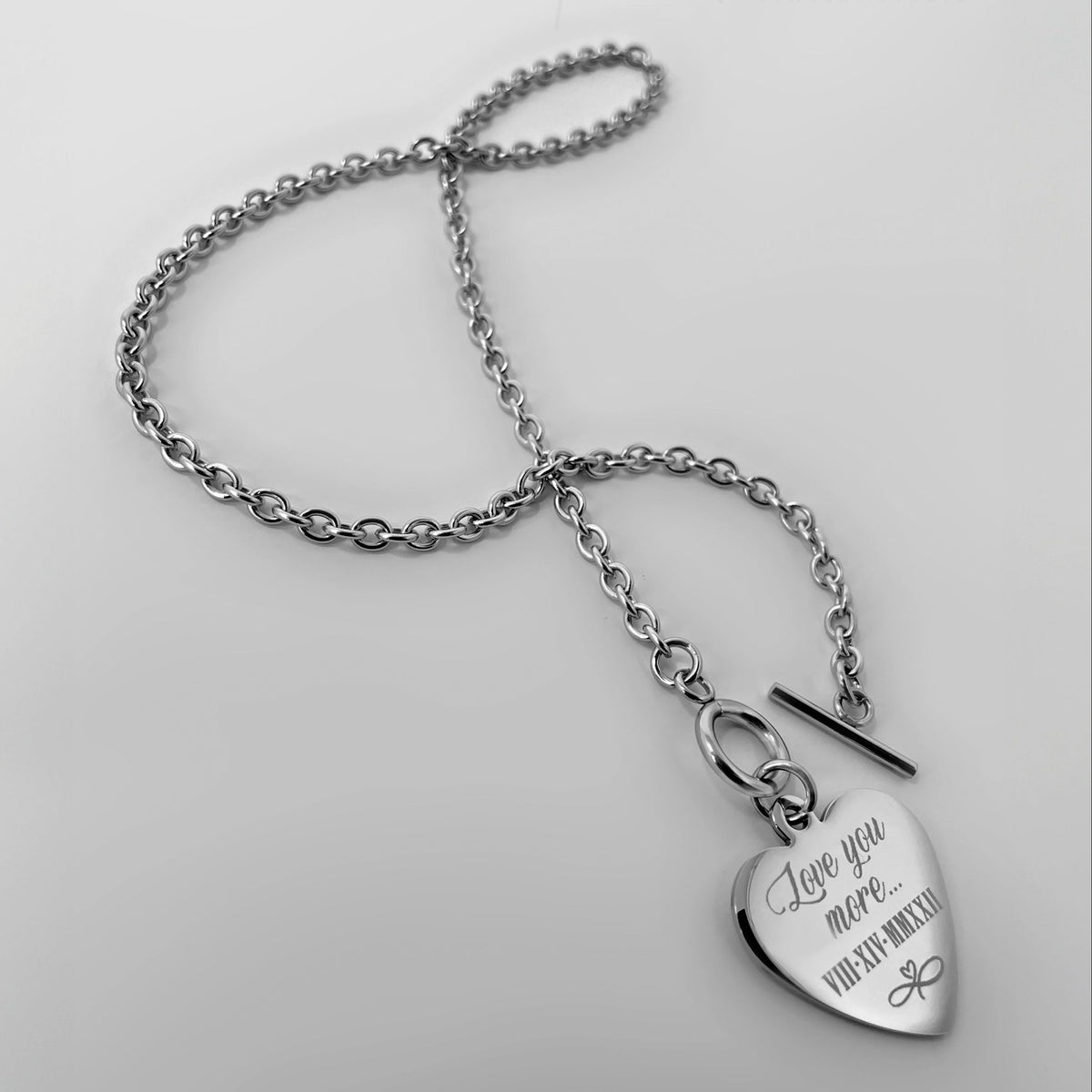 A silver toggle clasp chain necklace with a heart-shaped pendant engraved with a personalized message and a date, written in Roman numerals.