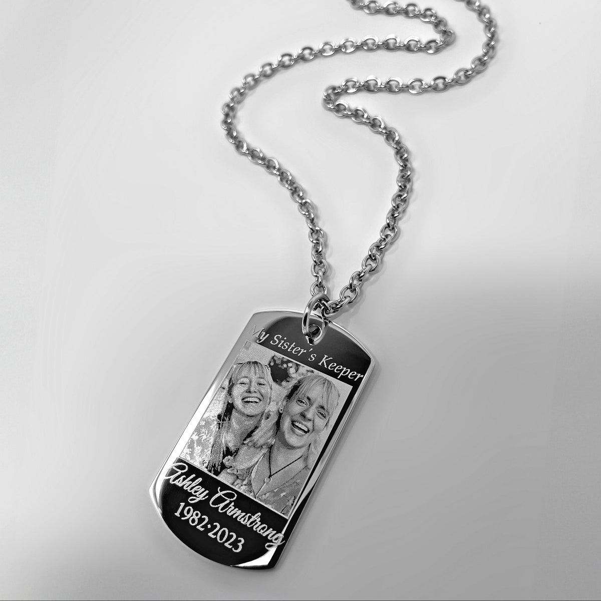 A silver necklace with a dog tag pendant engraved with a personalized photo of two sisters and memorial words 'My Sister's Keeper' and custom dates, shown on a plain light background.