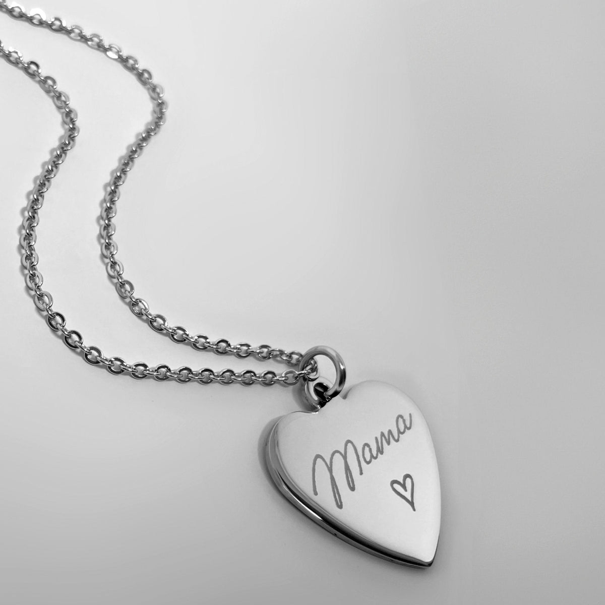 A silver heart-shaped pendant necklace engraved with the word 'Mama' and a small heart, shown on a matching silver chain against a light gray background.