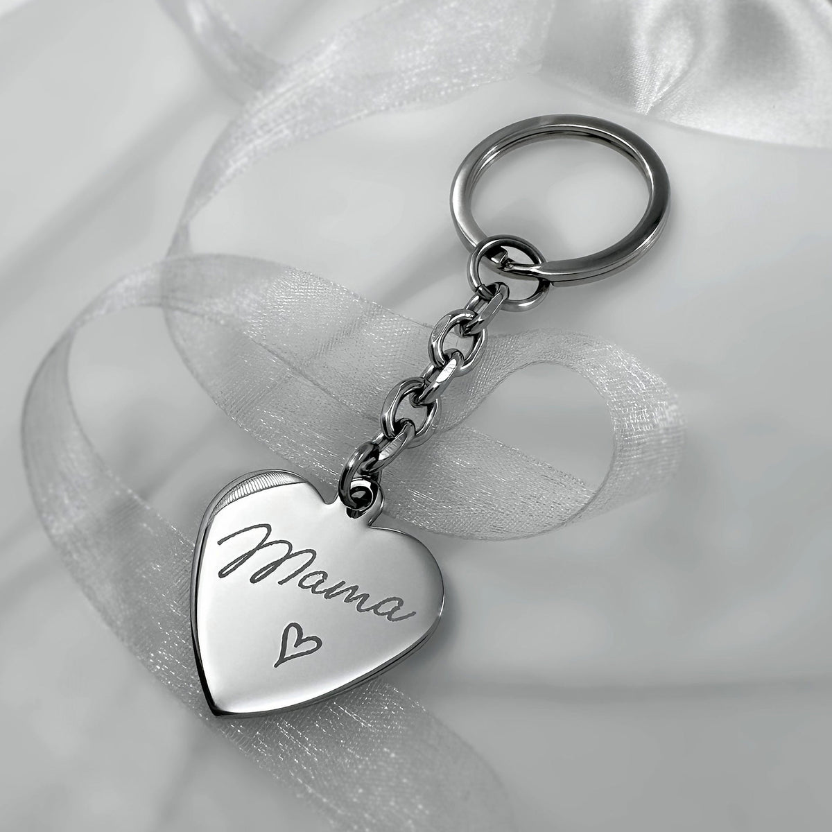 A silver heart-shaped keychain engraved with the word 'Mama' and a small heart, resting on a white satin ribbon.