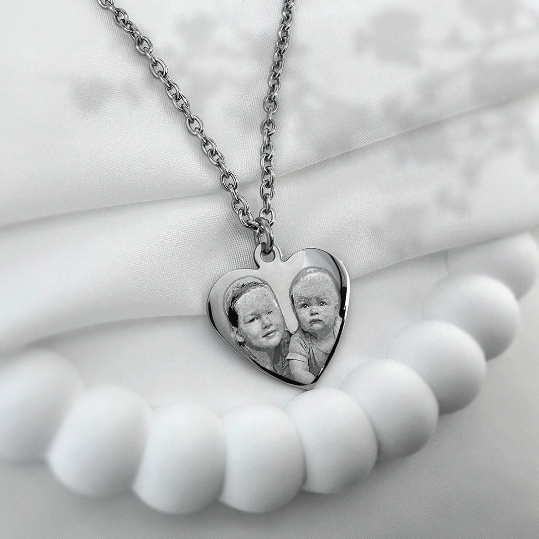 A silver heart-shaped pendant necklace featuring a custom photo of two children, displayed on a soft white fabric background.